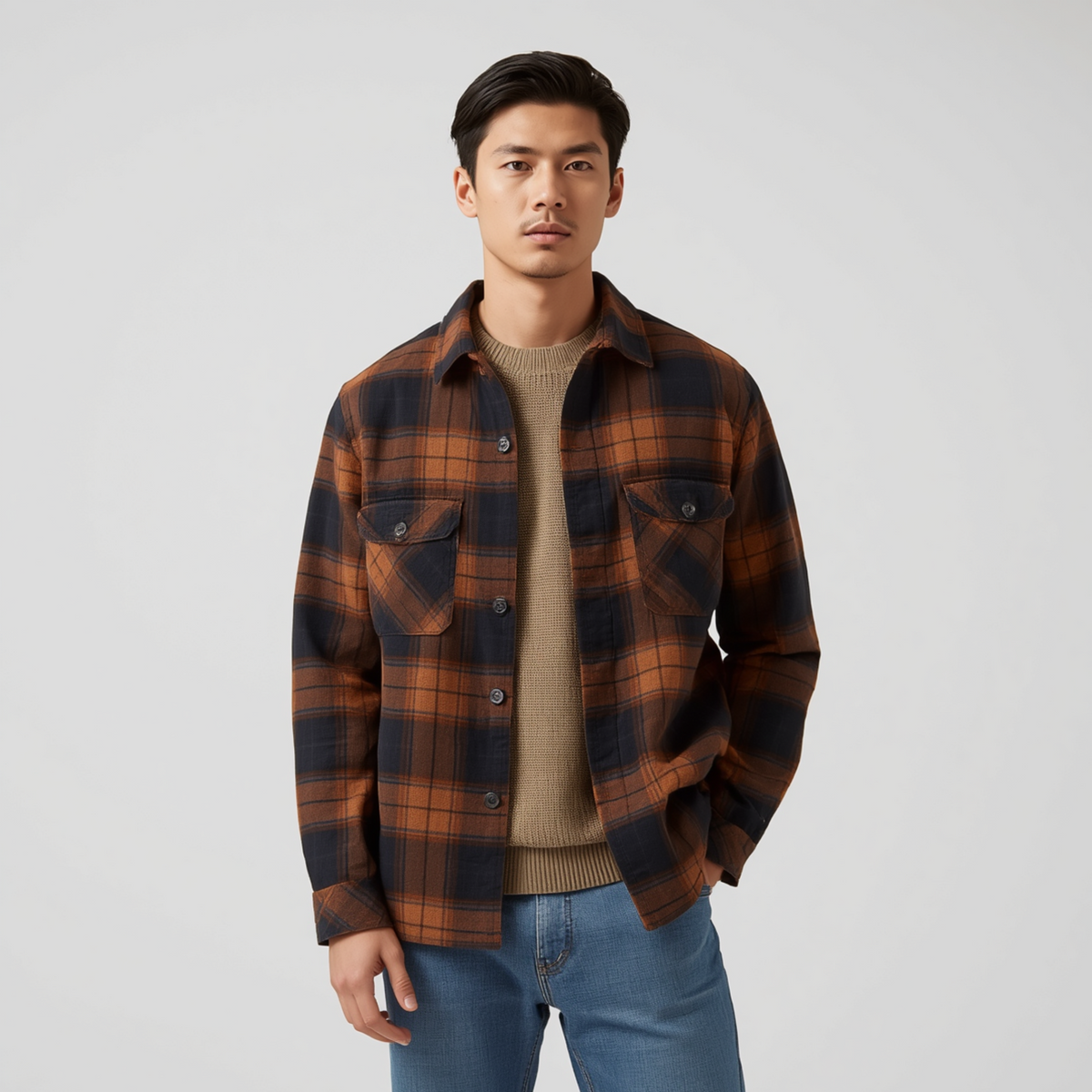 Mann trägt eine braune Karo-Hemdjacke mit Brusttaschen und Knopfleiste zu einem beigen Pullover und blauen Jeans vor hellem Hintergrund.