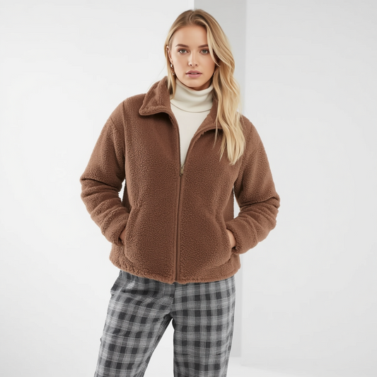 Frau trägt eine braune Teddy-Fleece-Jacke mit Reißverschluss und langen Ärmeln zu karierten Hosen vor hellem Hintergrund.   