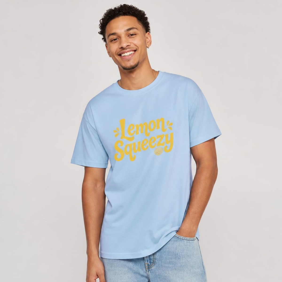 Mann trägt ein hellblaues T-Shirt mit gelbem "Lemon Squeezy"-Schriftzug zu hellen Jeans vor hellem Hintergrund.