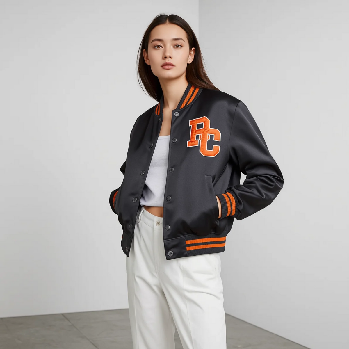 Frau trägt eine schwarze College Jacke mit orangefarbenem Aufnäher, Druckknöpfen und gestreiften Bündchen zu hellen Hosen vor hellem Hintergrund.