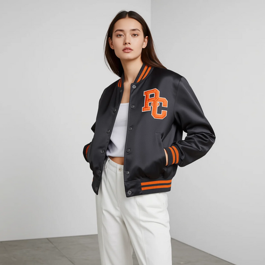Frau trägt eine schwarze College Jacke mit orangefarbenem Aufnäher, Druckknöpfen und gestreiften Bündchen zu hellen Hosen vor hellem Hintergrund.