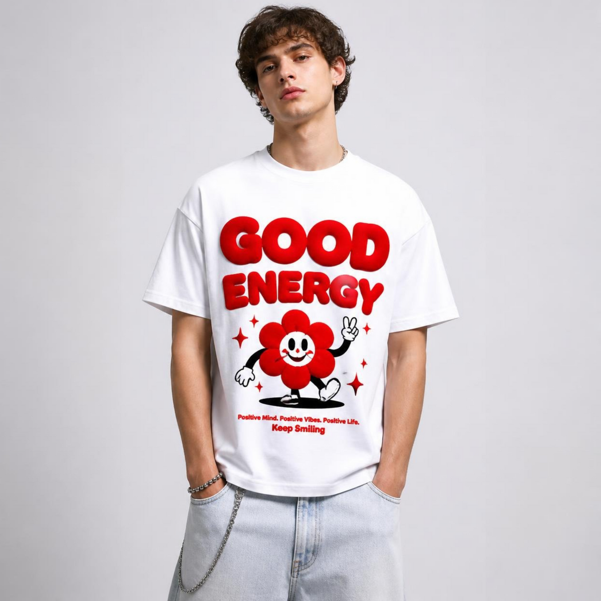 Mann trägt ein weißes T-Shirt mit rotem "Good Energy" Frontprint und Blumen-Grafik zu hellen Jeans vor hellem Hintergrund.   